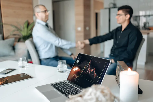 Two businessmen shaking hands in a modern office with trading data displayed on a laptop. Build a sponsorship rates reporting dashboard your clients trust